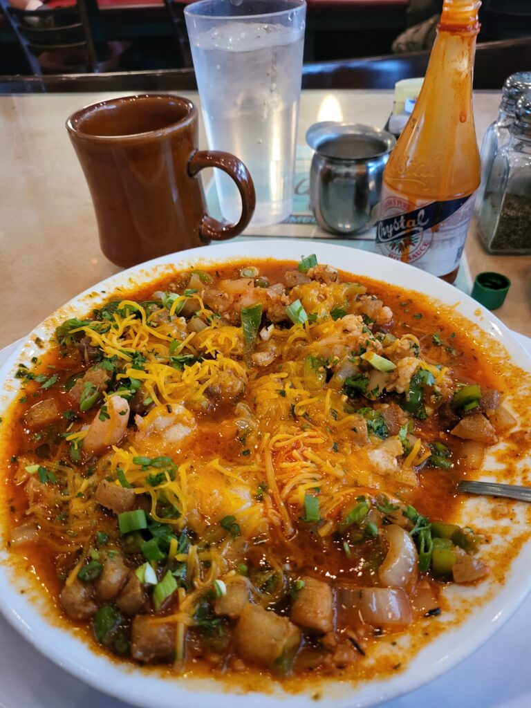 A plate of shrimp and grits with a cup of coffee. The food is bathed in a savory, spicy red sauce with chunks of onion and bell pepper and dressed with melted cheddar cheese.